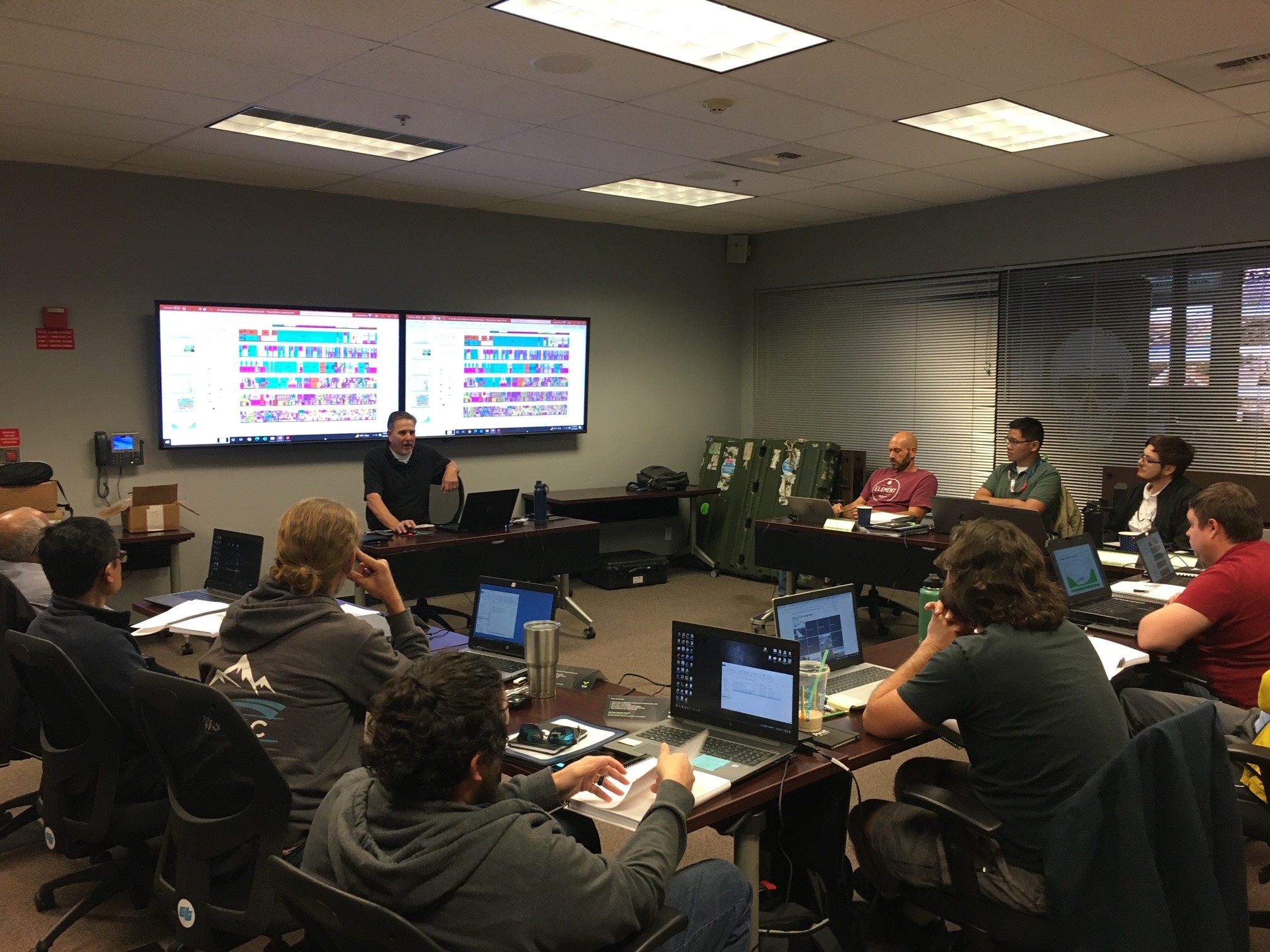 A group of people sit in a semi-circle around an instructor with a slide presentation up in the background.